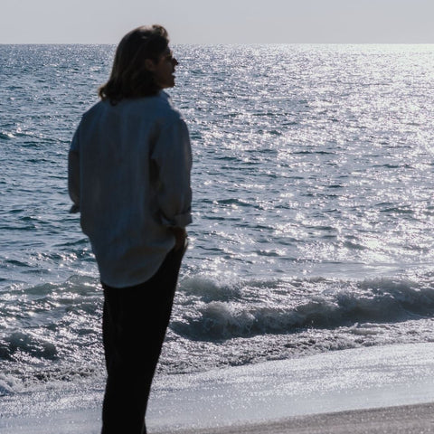A person stands on a beach shoreline facing the ocean, with waves rolling in and a pale sky above.